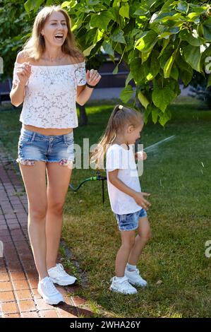 Mother with daughter walking under the water from the sprinkler irrigation in the summer city park. Childhood, leisure and people concept - happy fami Stock Photo