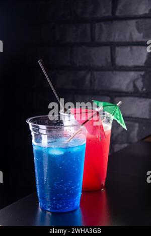 Red and blue lemonade with ice cubes in a plastic glasses decorated with an umbrella and straws on black wooden counter against a brick wall Stock Photo