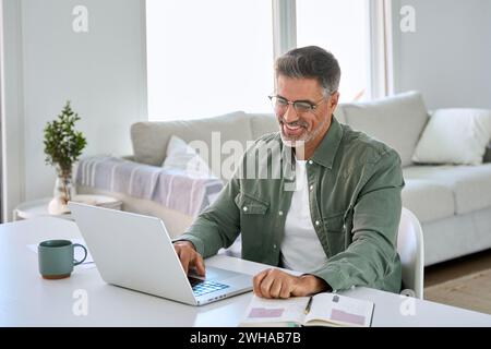Happy older middle aged man using laptop computer at home table. Stock Photo
