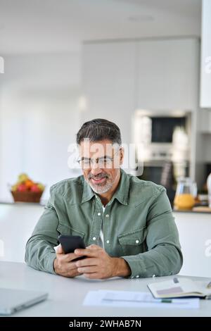Smiling middle aged man holding cell phone sitting at home table. Vertical Stock Photo