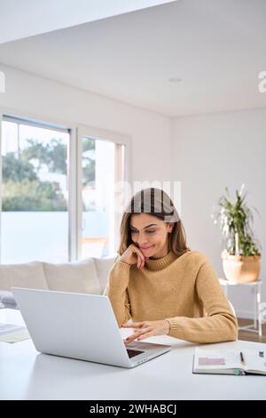 Smiling mature woman using laptop computer sitting at table in living room. Stock Photo