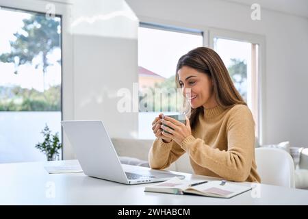 Smiling happy middle aged woman drinking coffee looking at computer at home. Stock Photo