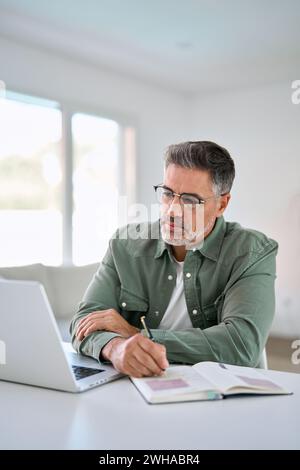 Serious older mature man using laptop computer writing notes at home table. Stock Photo
