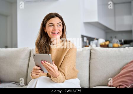 Happy mature woman using digital tablet sitting on couch at home looking away. Stock Photo