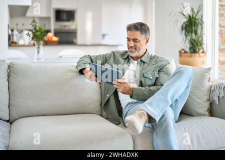 Happy middle aged man using digital tablet relaxing on couch at home. Stock Photo