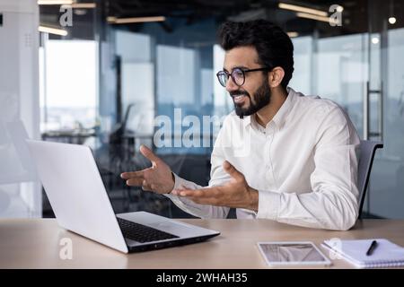 Online meeting, conference. Indian young man, office worker and businessman prays on laptop for video call, sitting in office at desk, gesturing with hands and smiling. Stock Photo