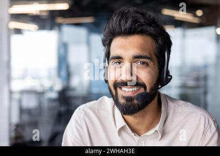 Portrait of a cheerful young Indian businessman with a headset, smiling in a bright, contemporary office workspace. Stock Photo