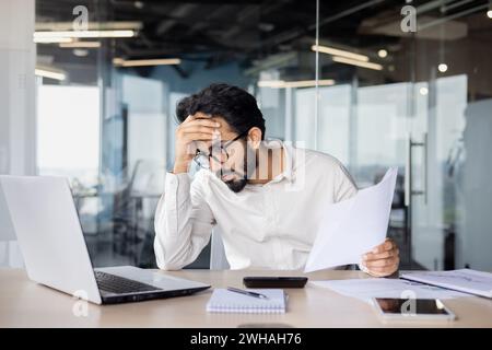 An Indian businessman with a beard deeply focused while working on his laptop at a modern office. He's reviewing documents, likely programming or strategizing. Stock Photo