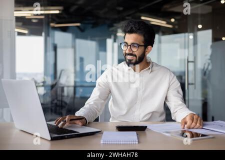 Confident Indian man with a beard, in shirt, programming on laptop at office desk. Exuding positivity and professionalism in a business environment. Stock Photo