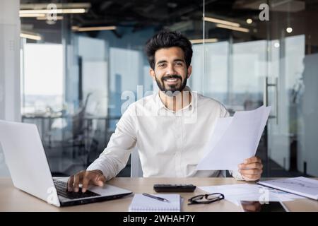 A man with a laptop is seen sitting at an engineering desk, holding a piece of paper and wearing a smile. Stock Photo