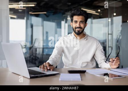 A white-collar worker is sitting at a desk with a laptop and wearing glasses, working on an engineering project with office equipment. Stock Photo