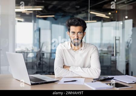 Serious Indian businessman in shirt with beard at office desk, programming on laptop with documents, experienced and focused. Stock Photo