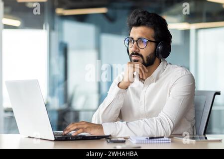 A bearded man seated at a desk, wearing glasses, is using a laptop computer and vision care eyewear. Stock Photo