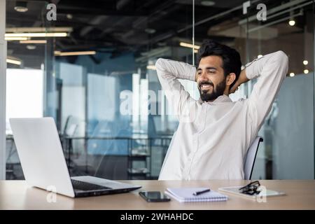 Content Indian businessman at his desk in a modern office, smiling as he takes a break from developing software on his laptop. Stock Photo