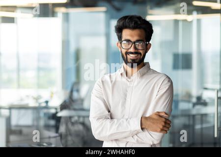 A man wearing formal attire, including a dress shirt and glasses, stands in an office with his arms crossed and a smile on his face. Stock Photo