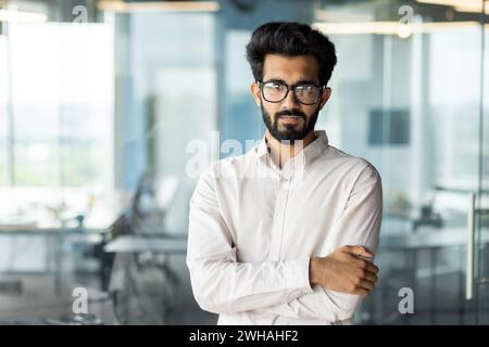 A white-collar worker, wearing a dress shirt, with a beard and glasses, is standing in an office, arms crossed, wearing a smile. Stock Photo