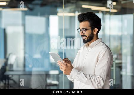 A man wearing a hat and beard stands by a glass wall, holding a tablet computer in his sleeve, displaying content using his thumb gesture. Stock Photo