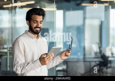 Smiling bearded Indian man using a tablet in a well-lit modern office. Portraying a tech-savvy businessman or a software developer at work. Stock Photo