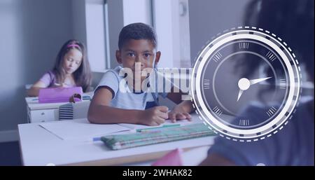 Image of clock over smiling biracial boy sitting at desk in diverse class Stock Photo