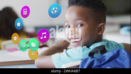 Image of school icons over happy african american schoolboy sitting at desk in class Stock Photo