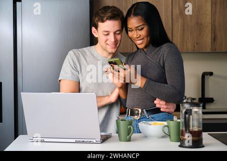 Multiethnic couple using the phone while having breakfast at kitchen. Stock Photo