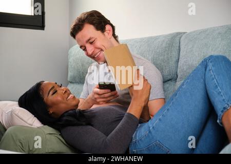 Multiethnic couple reading a book while rest in a sofa at home. Stock Photo