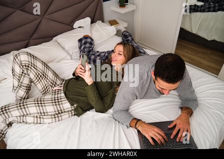Spanish couple using the laptop and the smartphone in bed. Stock Photo