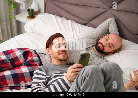 Homosexual couple using the phone together relaxing in bed. Stock Photo