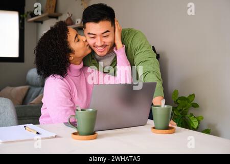 Multiethnic happy couple kissing on a cheek while using the laptop. Stock Photo
