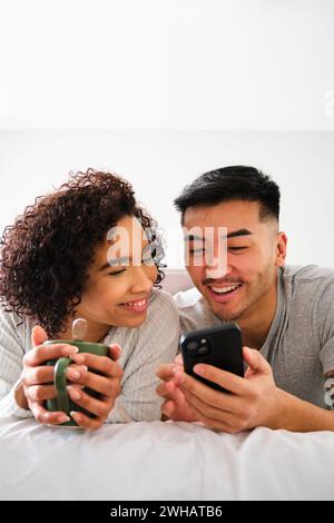 Multiethnic happy couple using the smartphone on bed. Stock Photo