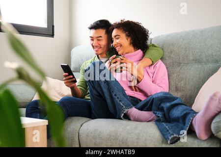 Multiethnic happy couple using the smartphone together on sofa. Stock Photo