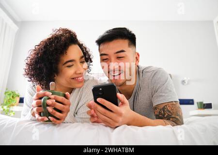 Multiracial happy couple using the smartphone on bed. Stock Photo