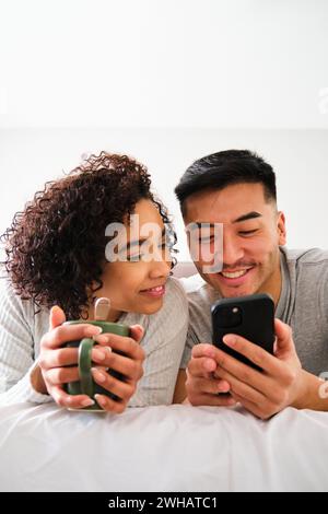 Multiracial couple smiling and using the phone on bed. Stock Photo