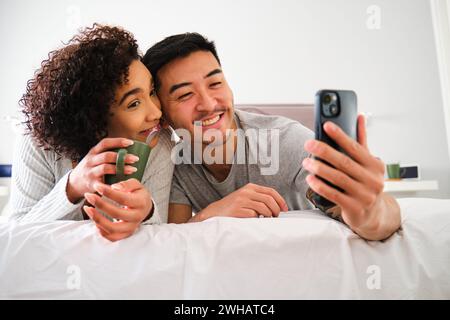 Multiracial couple smiling and taking a selfie on bed. Stock Photo