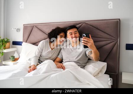 Multiethnic couple smiling and taking a selfie on bed. Stock Photo