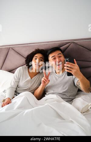 Diverse couple smiling and taking a selfie on bed. Stock Photo