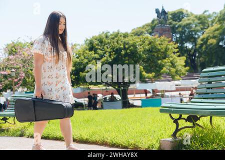 young latin woman street artist wearing white dress, walking in square with violin case to make music on street, copy space Stock Photo
