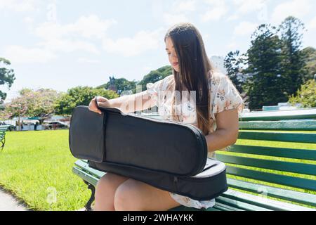 young Venezuelan Latin woman with long hair, wearing white dress, street artist is sitting on wooden bench in plaza taking her violin out of case to w Stock Photo