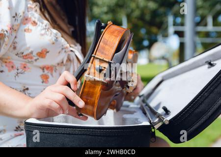 Close-up of unrecognizable Caucasian female artist sitting outdoors in park holding violin with her hands taking it out of case, preparing to play. Stock Photo