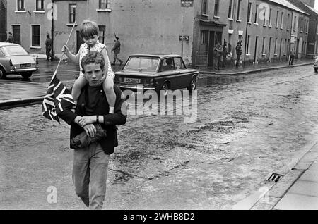 1970s Belfast. a young boy and his sister who is carrying a Union Jack flag return from an Orange Day parade. Walking through depressed back streets, while the British army patrol this inner city area. Northern Ireland. 1970 HOMER SYKES Stock Photo