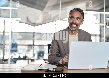 Smiling middle aged business man sitting at desk using laptop looking away. Stock Photo