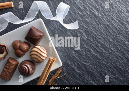 Assortment of chocolate bonbons on white ceramic plate on a slate table with white ribbon and cinnamon sticks. Top view. Stock Photo