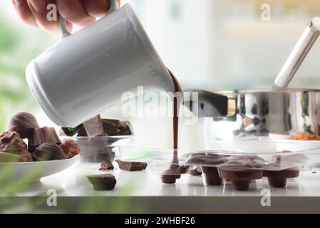 Person making chocolate bonbons on white kitchen bench at home with pouring hot chocolate into transparent mold. Front view. Stock Photo
