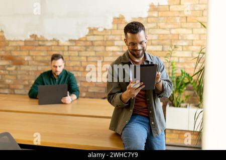 In a contemporary office setting, two focused colleagues engage in their work with tablet and laptop, collaborating and sharing ideas in a bright and Stock Photo