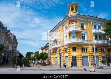 A yellow hotel building under a light blue sky with few clouds and passers-by on the street, hotel in the pedestrian zone Alexandrovska, Russe Stock Photo