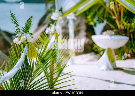 Decorated romantic wedding celebration accessories on tropical sandy beach. Lush green foliage and white lowers. Stock Photo