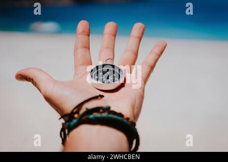 Open palm with stretched fingers holding black metal compass against white sandy beach. Find your way or goal concept. Point of view pov. Stock Photo