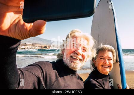 couple of pensioners seniors taking a selfie together at the beach with their wetsuits and surfboards - mature people learning surfing Stock Photo