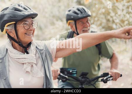 Two happy old mature people enjoying and riding bikes together to be fit and healthy outdoors. Active seniors having fun training in nature. Portrait of one old man smiling in a bike trip with his wife. Woman indicating something and looking at it. Stock Photo