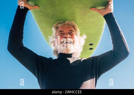 happy senior with surftable on his head is smiling and laughing - old and mature man having fun surfing with a black wetsuits - active retired adult doing activity alone Stock Photo